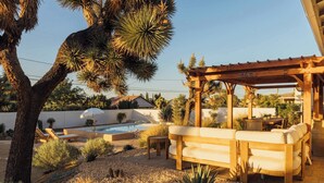 A patio with a wooden pergola, a pool, and a seating area with a sofa and chairs. - Hidden Gold House | Luxury Joshua Tree Retreat (Yucca Valley)