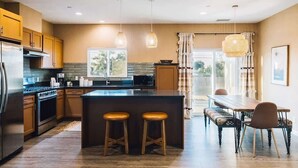 A modern kitchen with wooden cabinets, a central island with stools, a dining table with chairs, and pendant lighting. - Hidden Gold House | Luxury Joshua Tree Retreat (Yucca Valley)