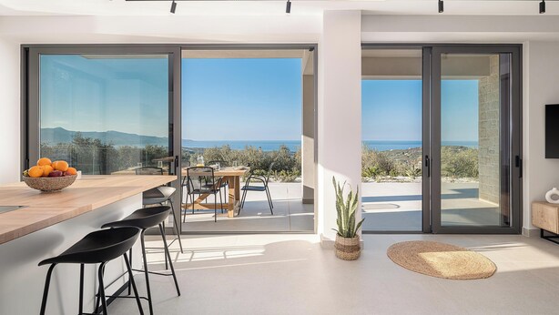 A modern kitchen with a large island, bar stools, and a view of the sea. - Villa Zefyros With Sea View (Platanias)