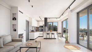 A modern kitchen with a central island, a dining area with a table and chairs, and a large sliding glass door leading to an outdoor patio. - Villa Zefyros With Sea View (Platanias)