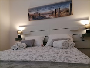 Ein ordentlich bezogenes Bett mit aufgerollten Handtüchern, ein Kopfteil mit einem Strandbild und zwei Nachttischlampen.