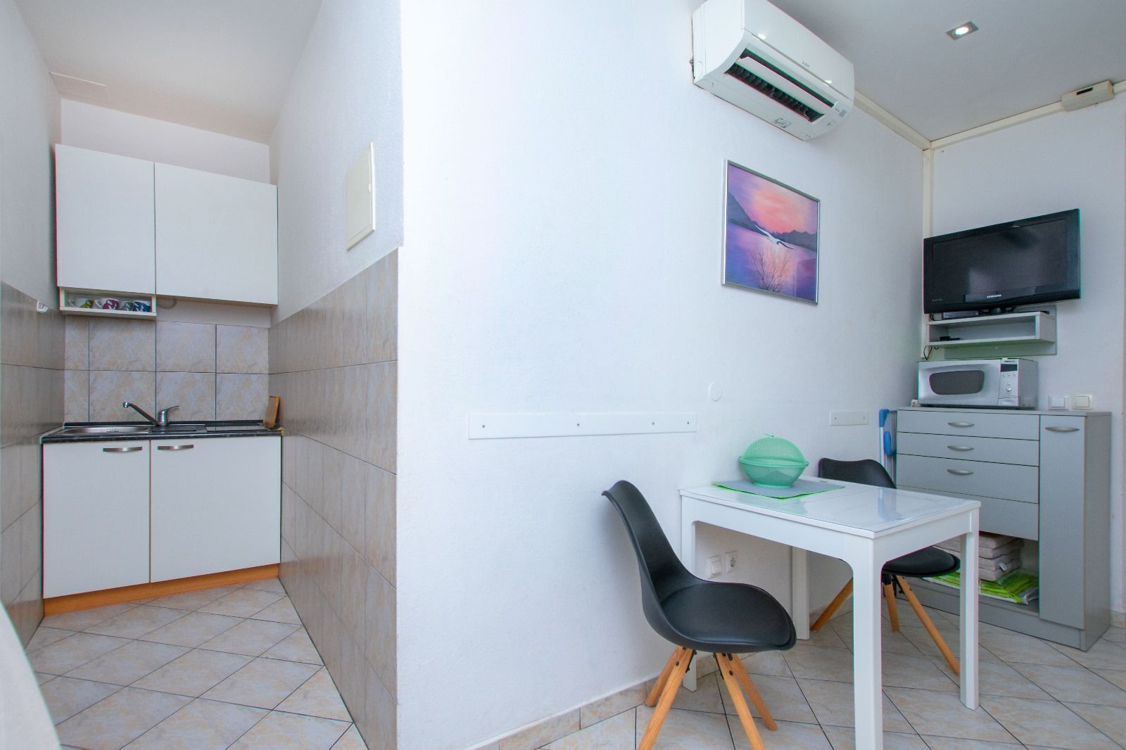 A compact kitchenette with a sink, cabinets, and an air conditioner. There is a small dining table with a chair and a TV mounted on the wall.
