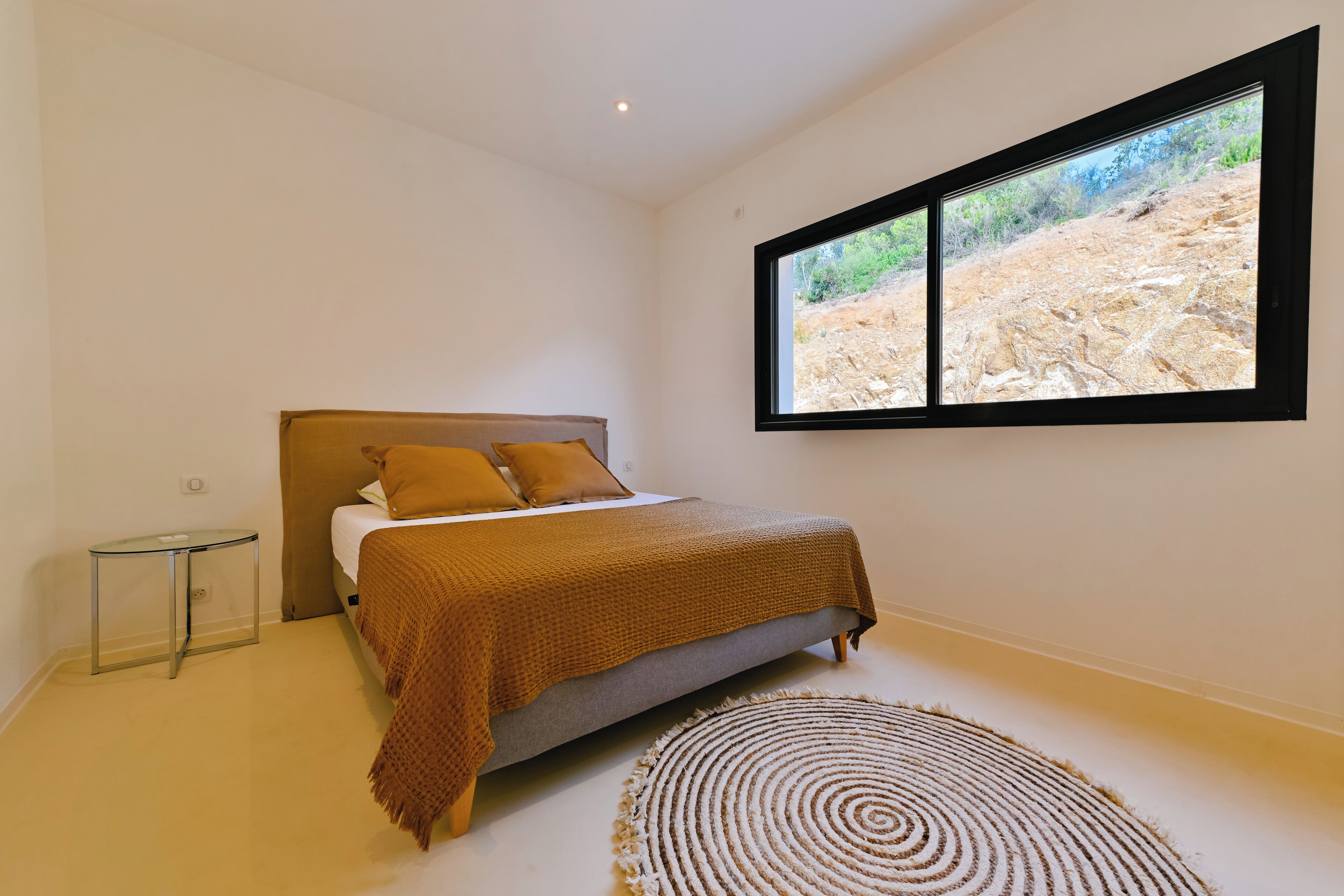 A bedroom with a bed, a small table, a window with a view of a rocky landscape, and a circular rug.