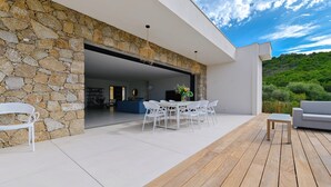 A modern outdoor patio with a stone wall, a white table and chairs, and a wooden deck. - SUMPTUOUS MODERN VILLA (Pietrosella)