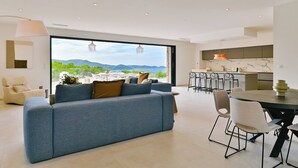 A modern living room with a large sofa, dining table, and chairs, a kitchen area, and a large window with a view of mountains. - SUMPTUOUS MODERN VILLA (Pietrosella)