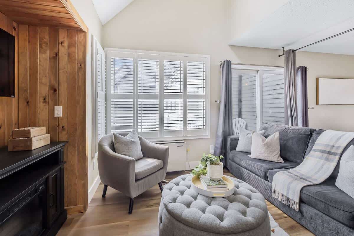 A living room with a grey sofa, a grey armchair, a wooden TV stand, and a window with blinds.