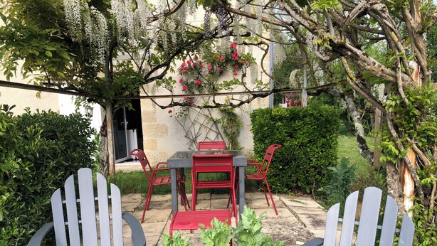 Eine Terrasse im Freien mit Tisch und Stühlen, umgeben von Grünpflanzen und einer Pergola mit hängenden Blumen.