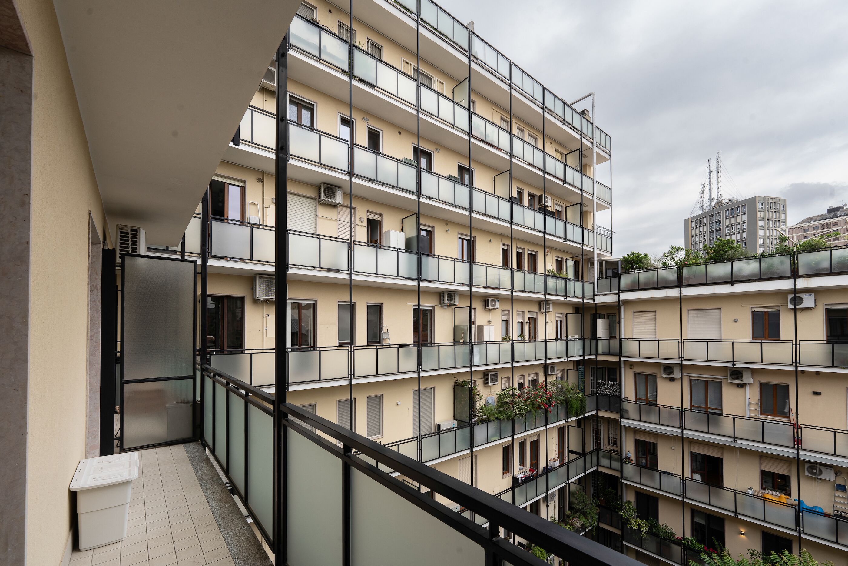 A multi-story residential building with balconies, air conditioning units, and a view of other buildings in the background.