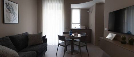 Un salon moderne comprenant un canapé, une table à manger avec des chaises, un coin cuisine et un téléviseur à écran plat fixé au mur.