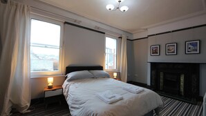 A hotel room with a bed, a nightstand with a lamp, a fireplace, and large windows with curtains.