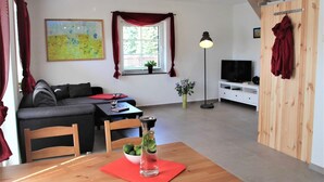 Un salon moderne avec un canapé, une table à manger agrémentée d’un set de table rouge, un grand vase en verre contenant une plante et un meuble pour la télévision.