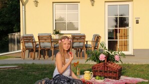 Une personne assise sur l’herbe, devant un pique-nique composé d’un panier, d’une carafe et d’un saladier rempli de fruits.