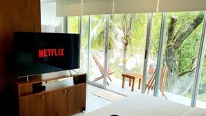 Et moderne hotelværelse med et stort fladskærms-tv, der viser 'NETFLIX', et træ-tv-skab, en pænt redt seng og udsigt til palmer gennem store vinduer.