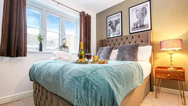 Ein Schlafzimmer mit einem großen Bett, einem Nachttisch mit Lampe, einem Fenster mit Vorhängen und zwei gerahmten Bildern an der Wand.