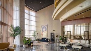 Eine großzügige Lobby mit hoher Decke, großen Fenstern und einem polierten Marmorboden. Mehrere Sitzbereiche mit Sofas und Sesseln sowie einige Kübelpflanzen sind vorhanden.