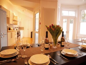 Une salle à manger bien éclairée, avec une table dressée pour quatre personnes, agrémentée d’assiettes blanches, de couverts et de verres à vin. Un bouquet de tulipes apporte une touche décorative.