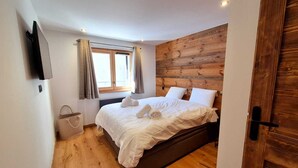 A bedroom with a wooden headboard, a bed with white linens, a window with curtains, and a wooden floor. - Sapins302 | 2024 Apt | Near center & slopes (Les Gets)