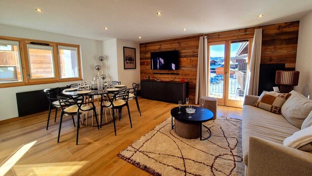 A modern living room with a dining area, wooden wall paneling, a flat-screen TV, and a view through a glass door. - Sapins302 | 2024 Apt | Near center & slopes (Les Gets)
