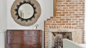 Une pièce avec une commode en bois, un miroir rond, une cheminée en briques et un tableau accroché au mur.