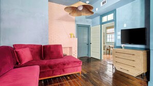 Une pièce avec un canapé rouge, un plancher en bois, un téléviseur posé sur une commode et une porte donnant sur une autre pièce.