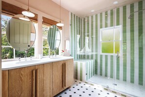 Une salle de bain avec un meuble-lavabo en bois, deux miroirs ronds, un lavabo blanc et une douche aux carreaux verts et blancs.