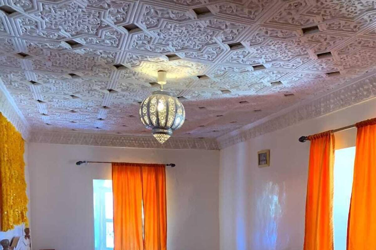 A room with a patterned carpet, a bed with a wooden frame, a nightstand, and a hanging lamp.