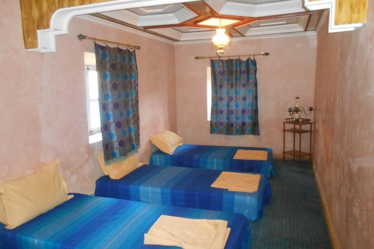 A room with three beds, each with blue bedding and a beige blanket. There is a small table with a vase and a lamp.
