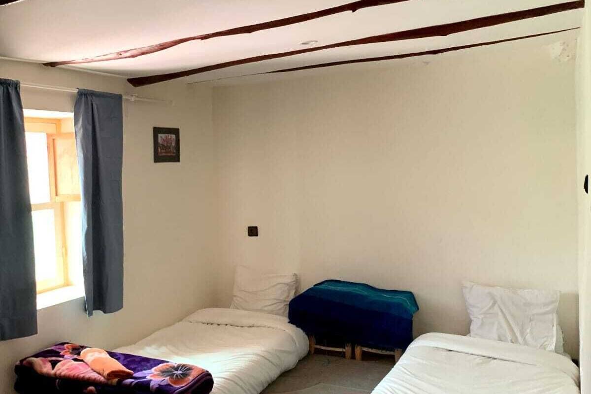 A small, cozy room with two single beds, a window with curtains, and a bed with a rolled-up blanket.