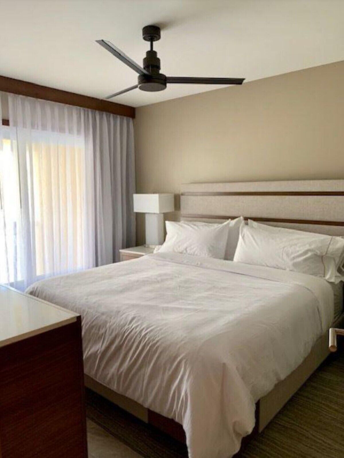 Quarto de hotel com cama, mesa de cabeceira, abajur e ventoinha de teto.