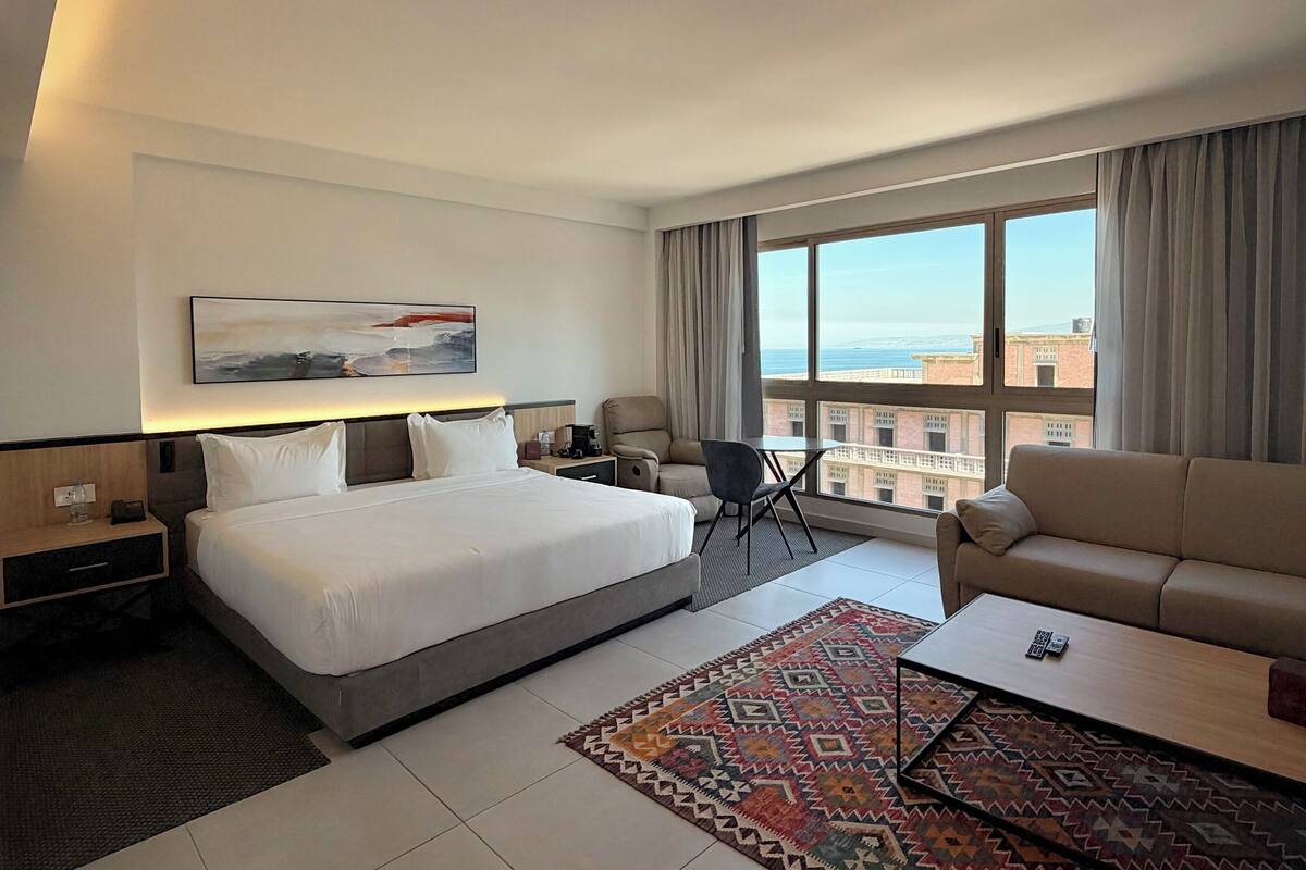 A modern hotel room with a large bed, a sofa, a dining table, and a view of the cityscape.