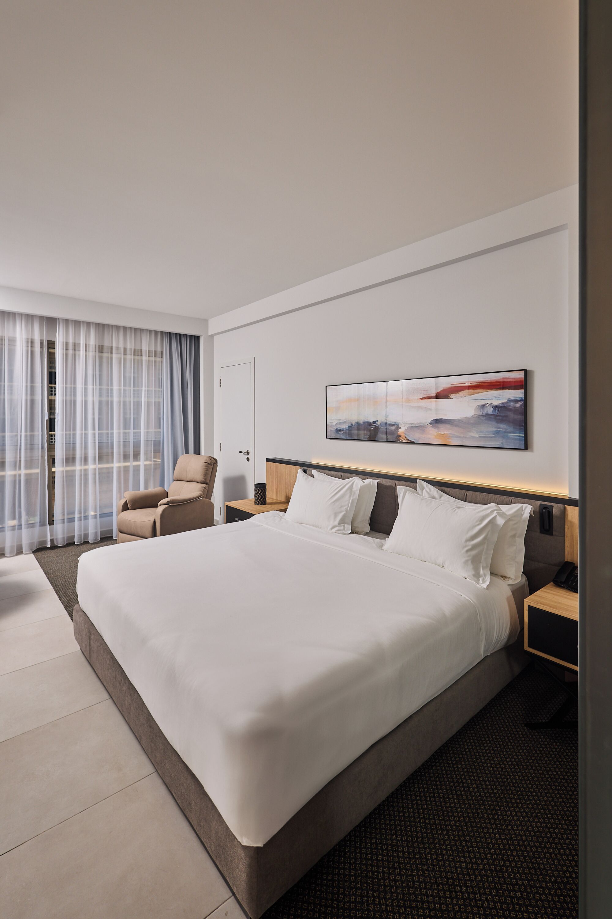 A hotel room with a large bed, a chair, a bedside table, and a wall-mounted artwork.