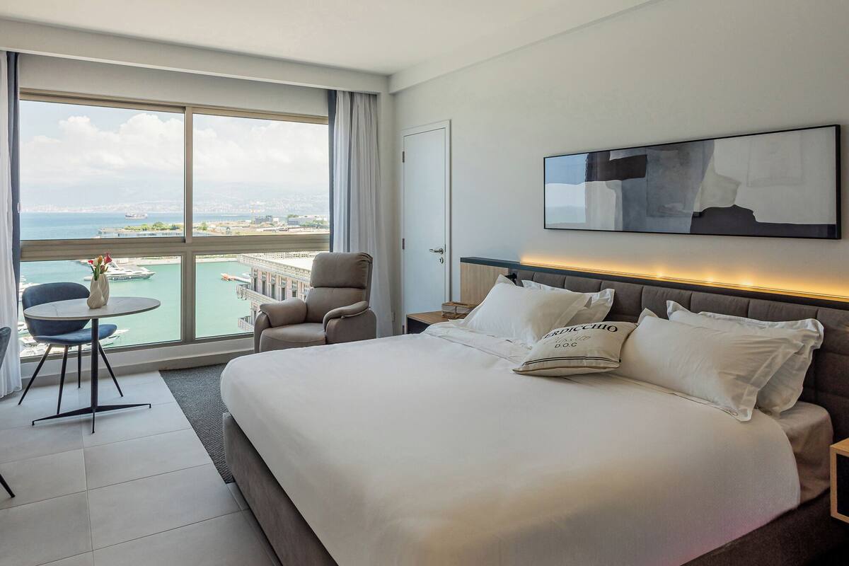 A modern hotel room with a large bed, a chair, a small table, and a view of the city and sea through large windows.