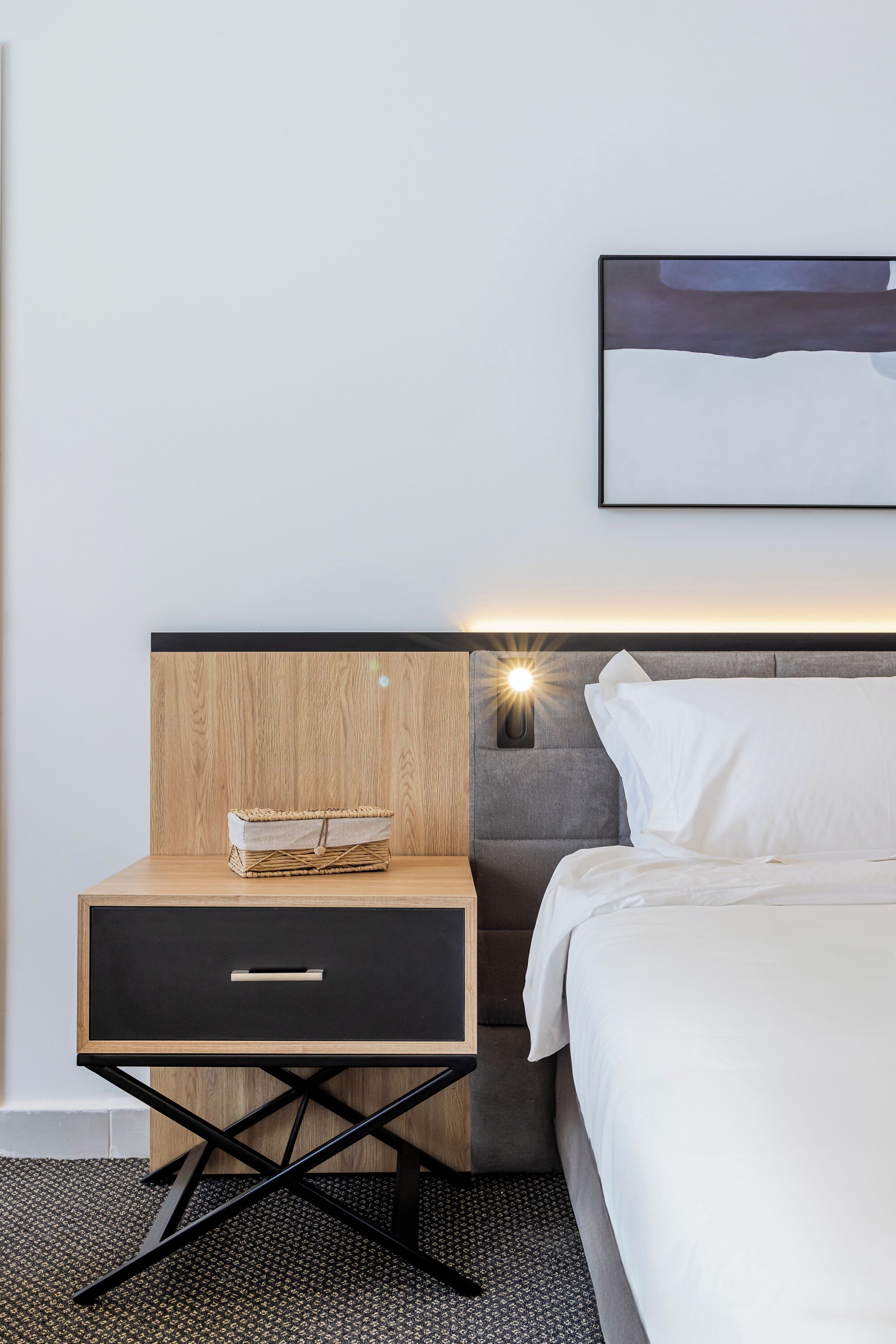 Una habitación de hotel moderna con una mesita de noche de madera, una pequeña cesta tejida, un cajón negro, una mesita con lámpara y una cama con ropa de cama blanca.