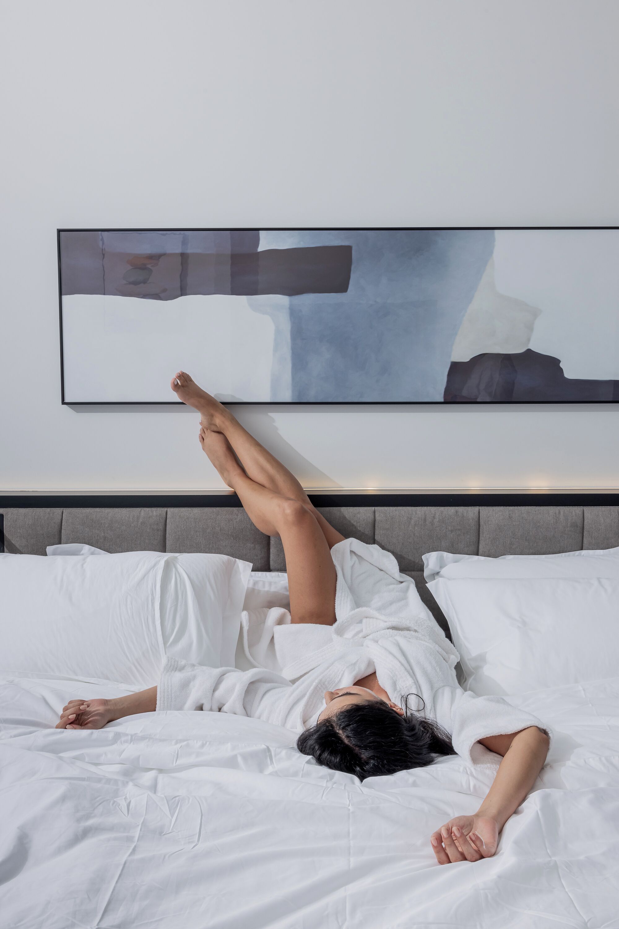 Una persona acostada en una cama en una habitación de hotel con ropa de cama blanca y un cabecero gris.