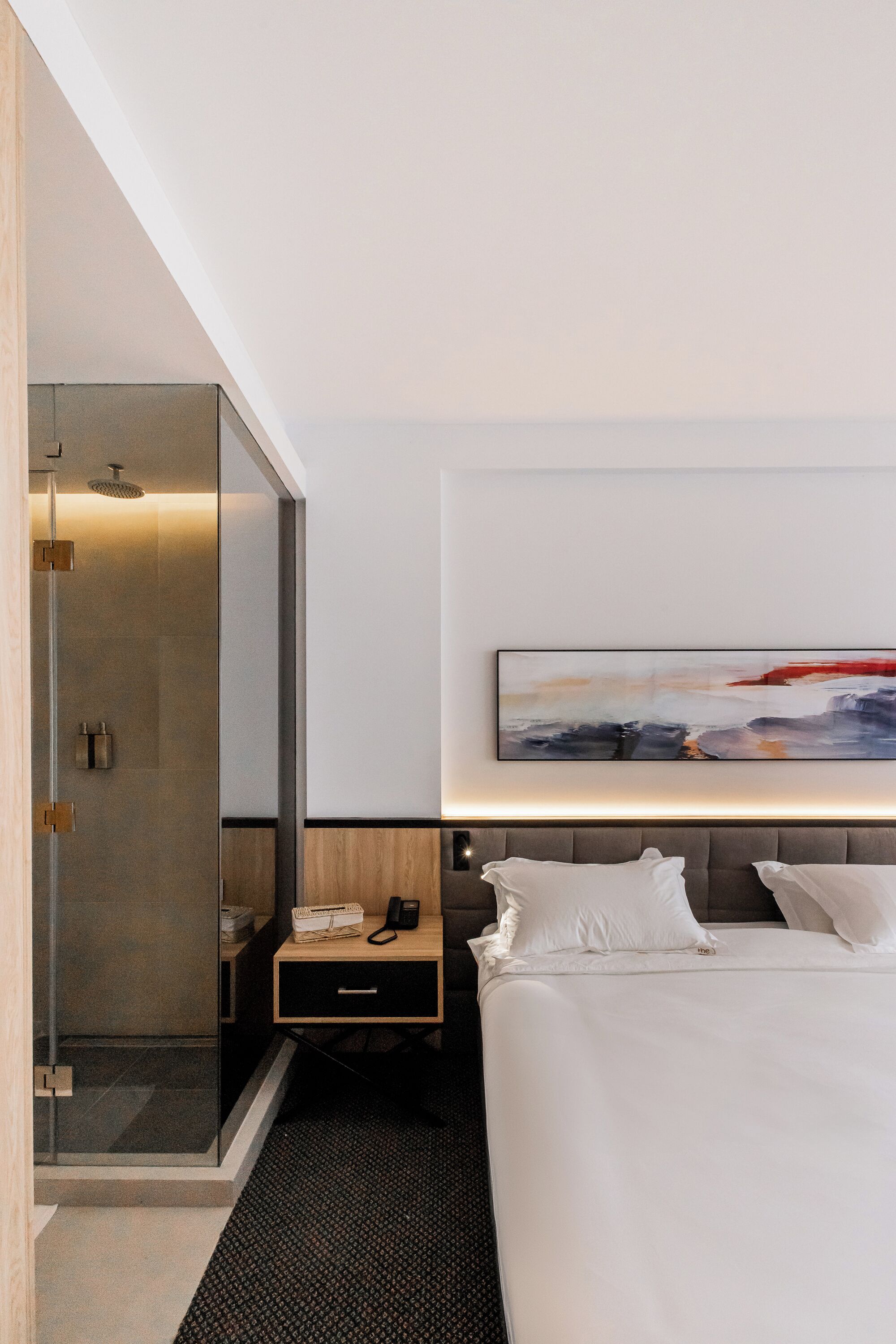 A modern hotel room with a glass-enclosed shower, a bedside table with a phone, and a bed with white linens.