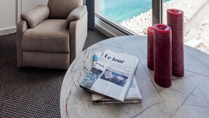 A room with a large window, a beige armchair, a round table with a newspaper, and two red candles. - The View - Saint Georges Hotel (Beirut)