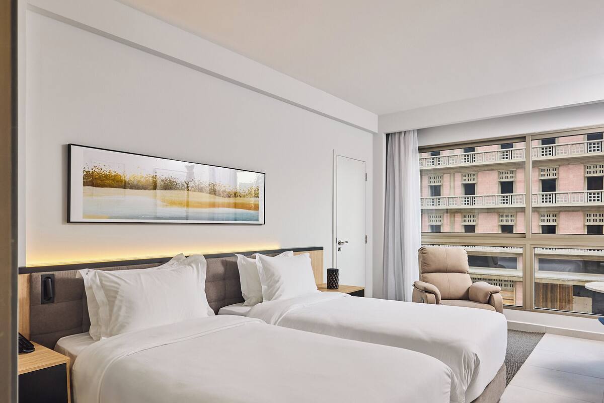 A hotel room with two beds, a large window with a balcony, and a painting on the wall.