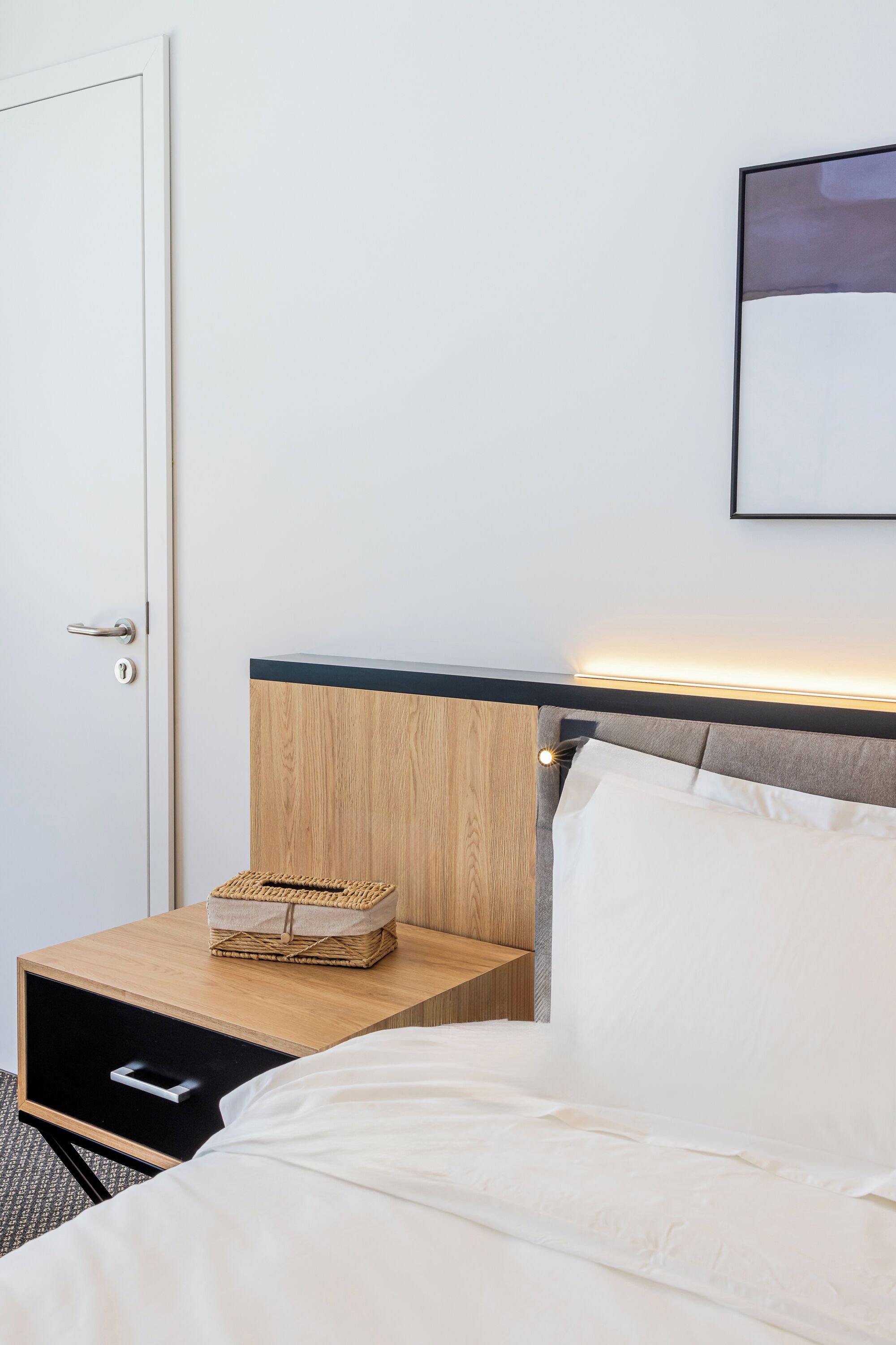A neatly made bed with white linens, a wooden nightstand with a basket, and a wall-mounted picture frame.