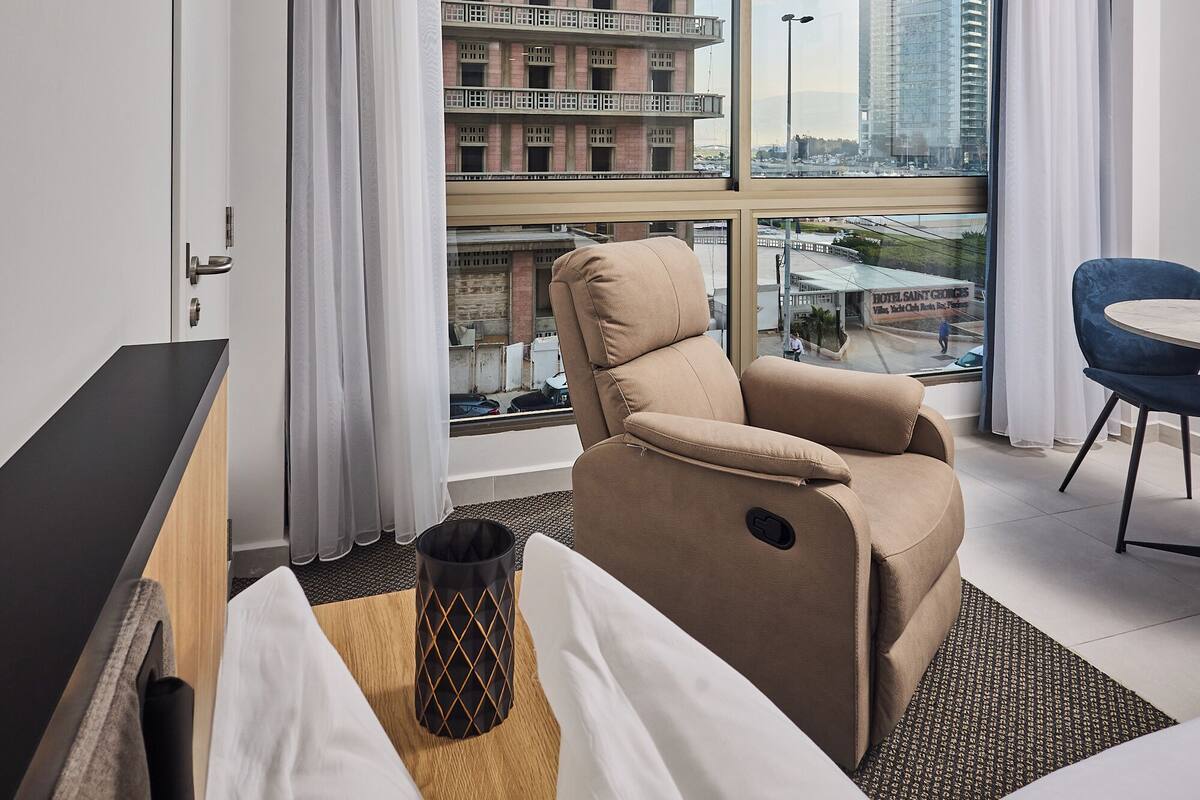 A modern hotel room with a large window, a sofa, a chair, and a bed.