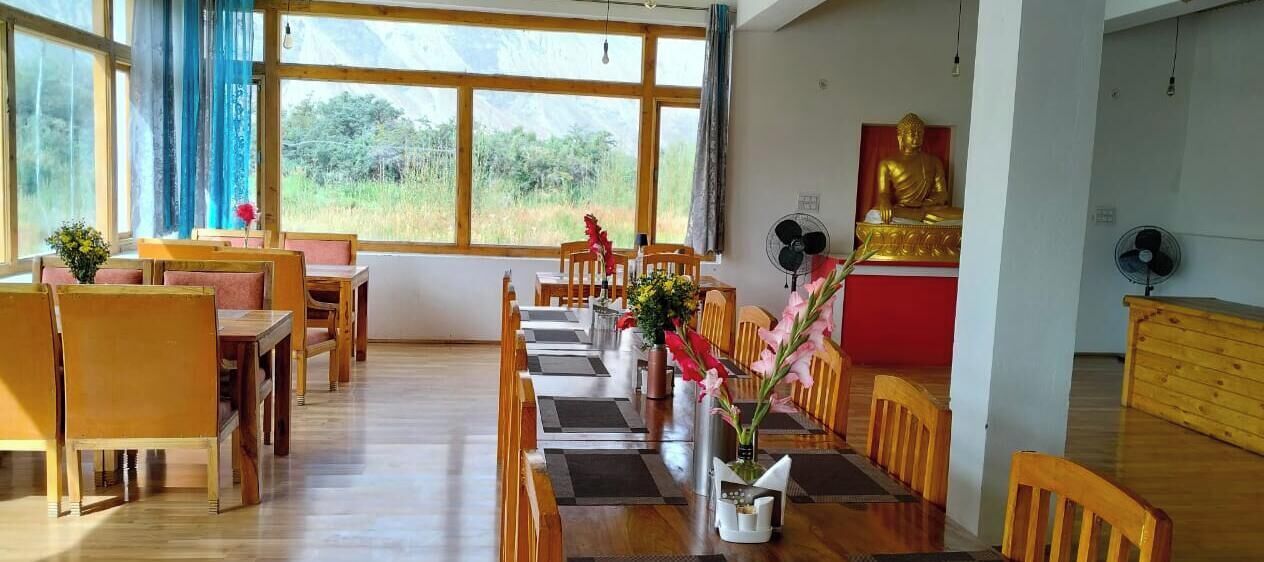 A dining area with wooden tables and chairs, a Buddha statue, and a view through large windows.