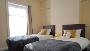 Ein Hotelzimmer mit zwei Betten, einem Fenster mit Vorhängen, einem Nachttisch mit Lampe und einer Kopfteil.