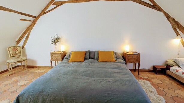A bedroom with a sloped ceiling, a large bed with a blue bedspread, two bedside tables with lamps, a chair, and a small table with a plant. - Two Berry houses with private garden and shared courtyard, perfect for groups (Thenay)