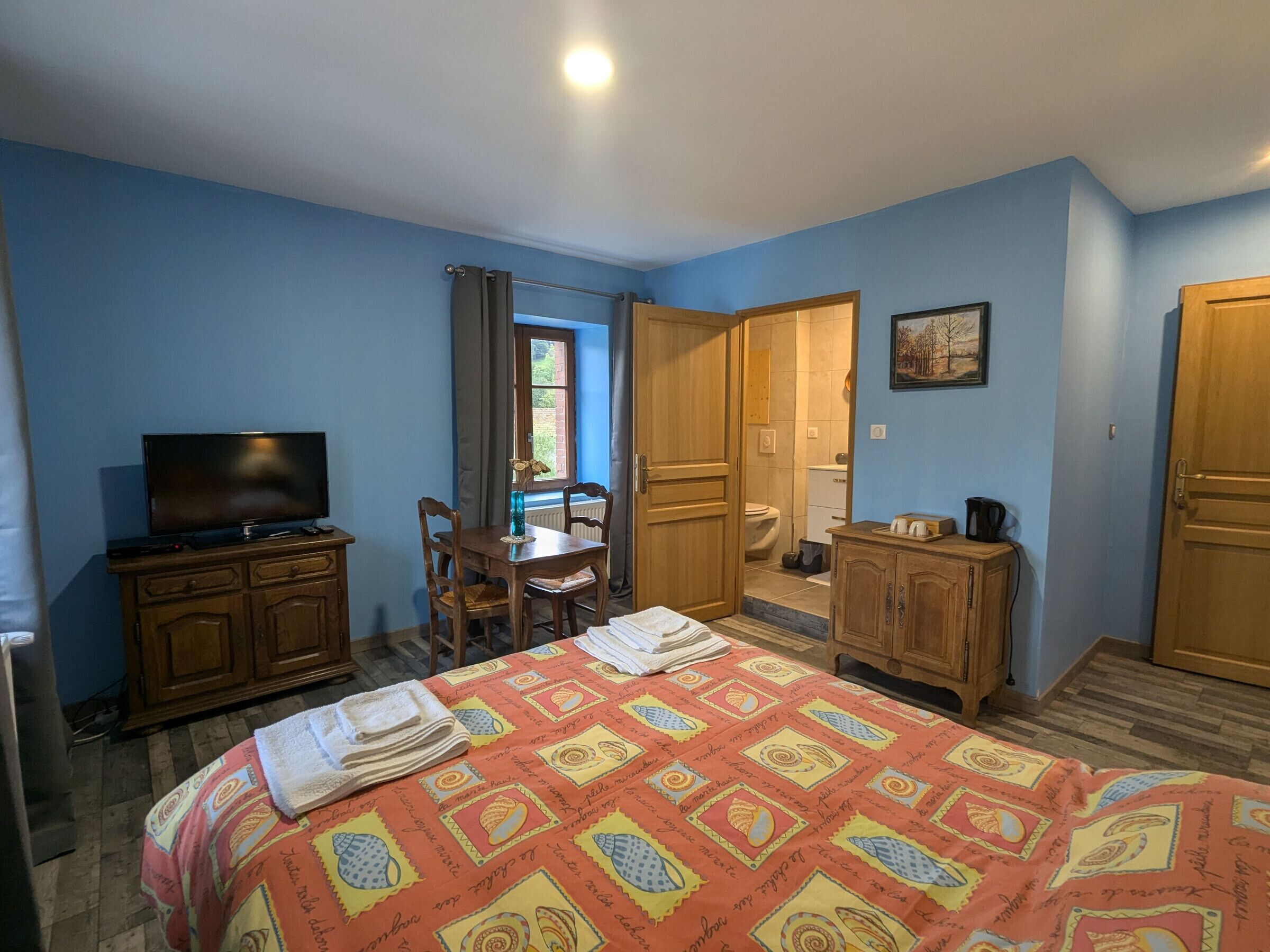 Een slaapkamer met een bed, een televisie, een houten dressoir, een tafel met stoelen, en een badkamer die door een open deur zichtbaar is.