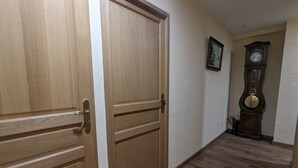 A hallway with wooden flooring and a large wooden door. A grandfather clock is visible in the background.