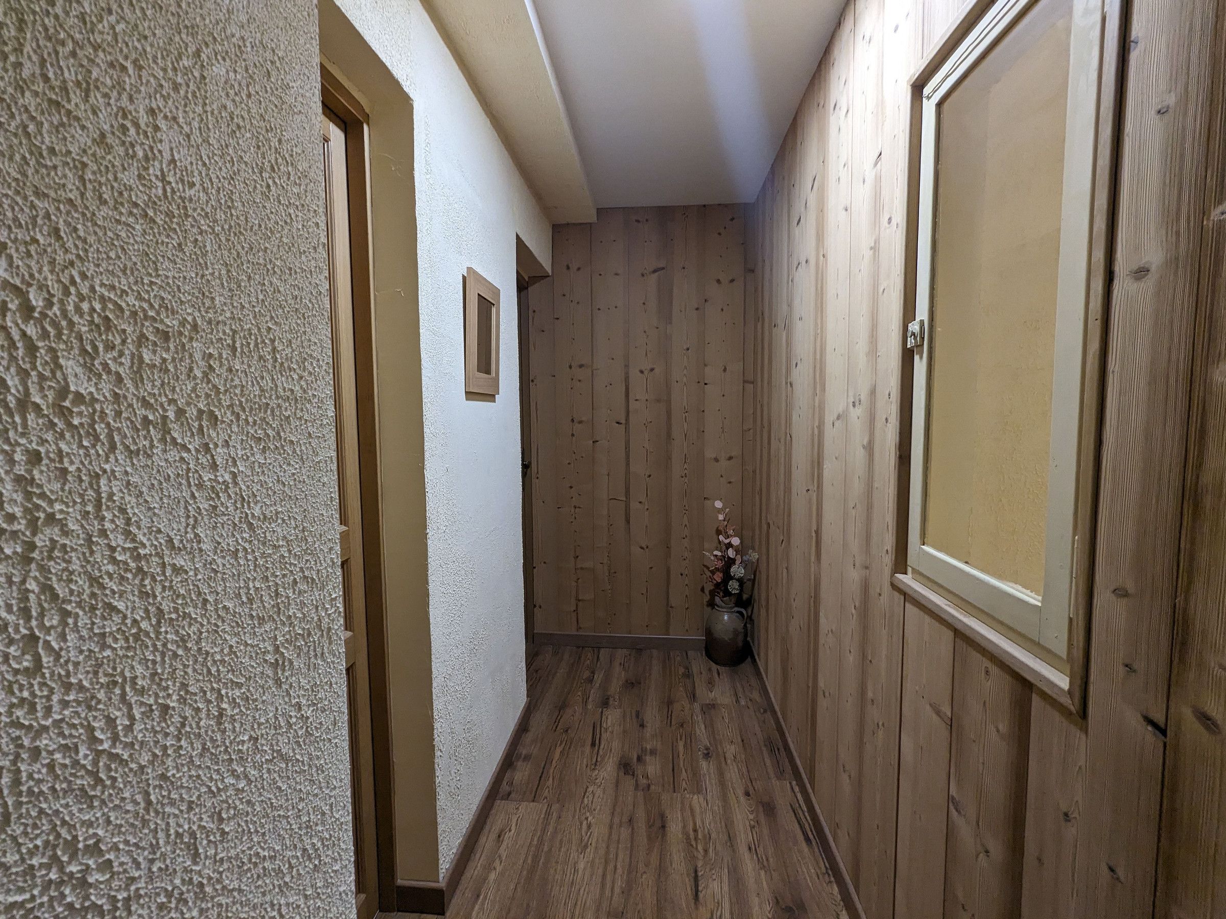 Ein schmaler Flur mit Holzwänden und Holzboden. Rechts befindet sich eine Tür und links eine strukturierte Wand.