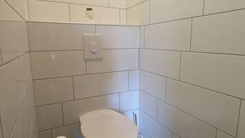 Un baño pequeño y limpio con azulejos blancos, un inodoro, un rollo de papel higiénico y una pequeña estantería con una planta.