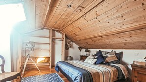 A cozy attic bedroom with a wooden ceiling, a bed with patterned bedding, a standing fan, and a chair.