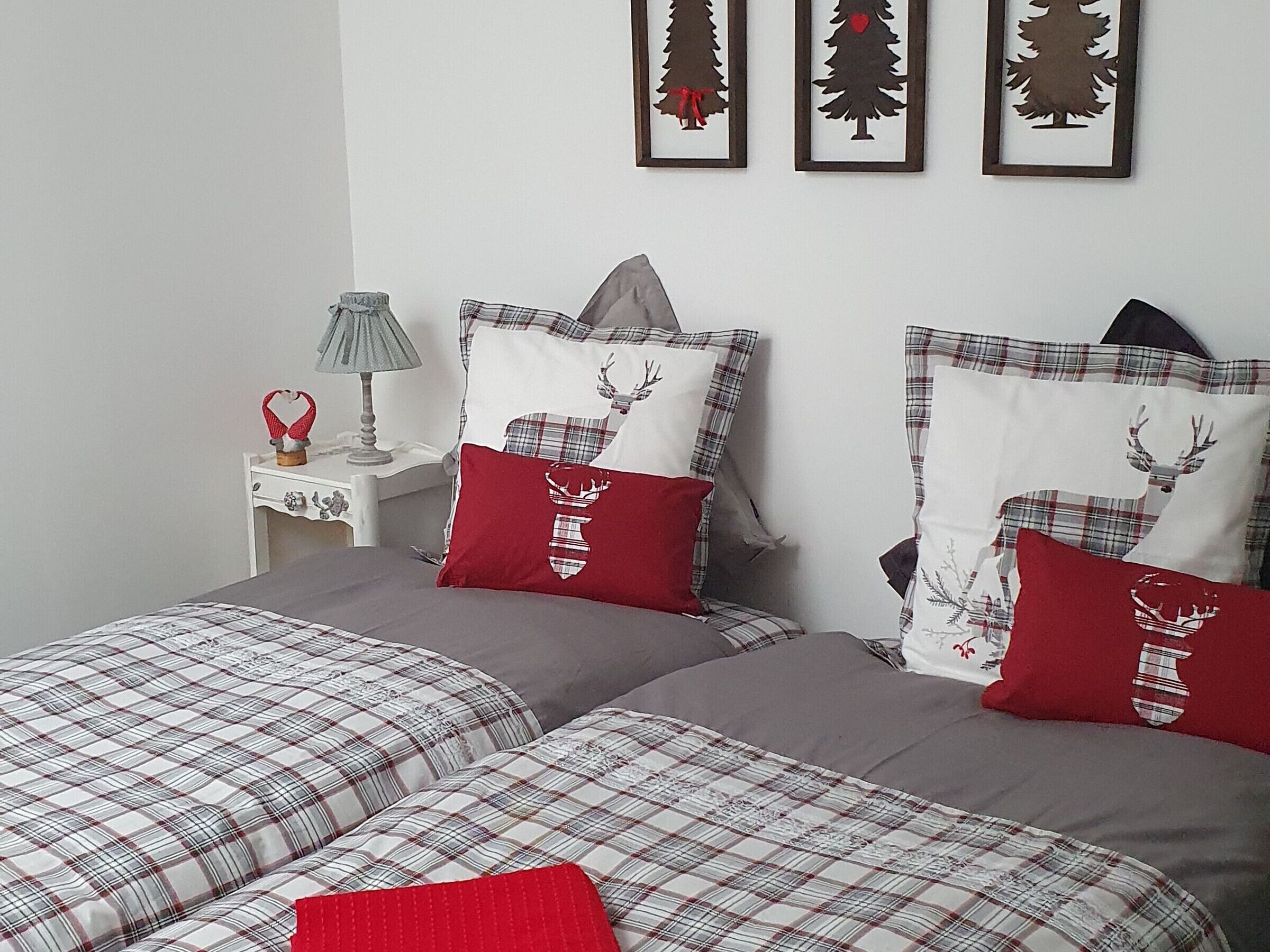 A bedroom with two beds, each with plaid bedding and red pillows, a nightstand with a lamp, and three framed Christmas tree decorations on the wall.