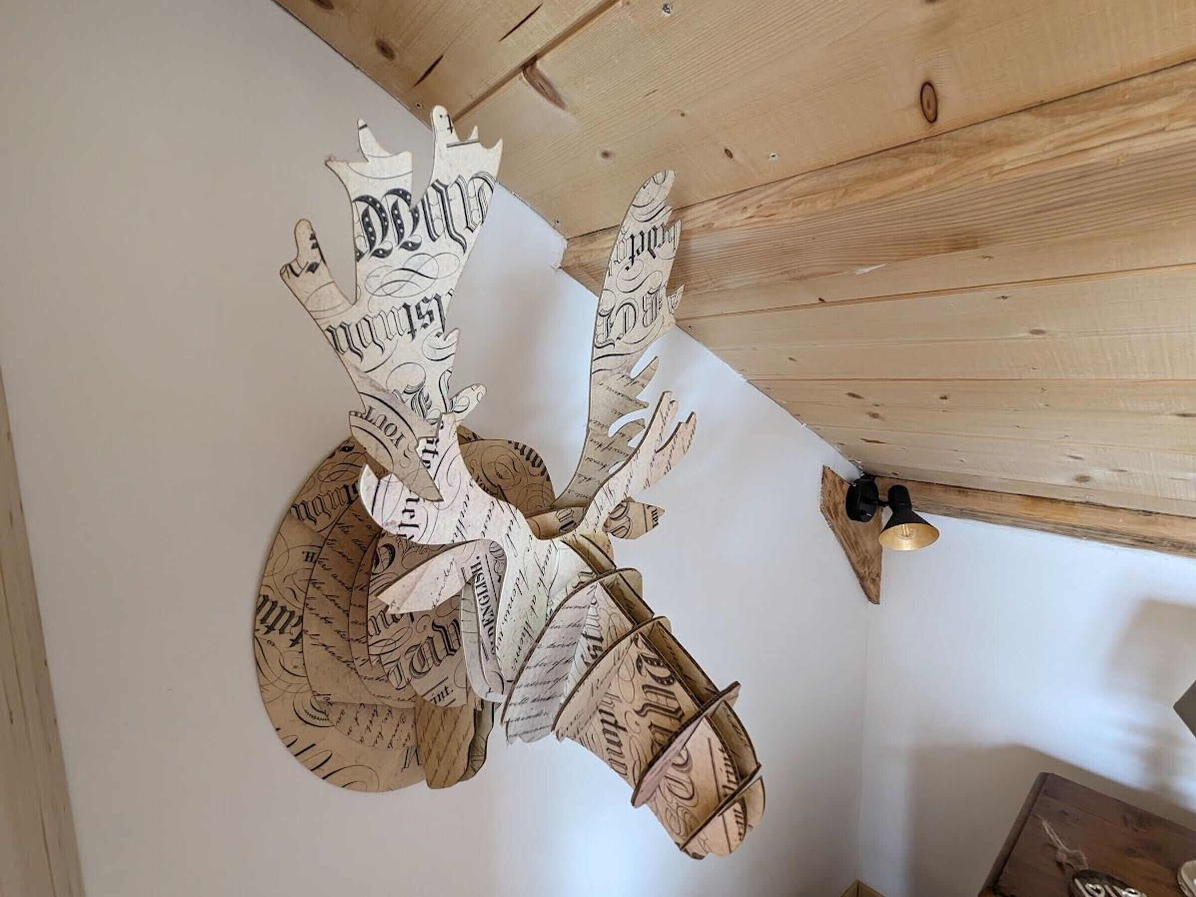 Un élément décoratif mural, fait de papier et portant un texte, peut-être une carte ou un document, dans une pièce aux murs lambrissés et comportant une petite table sur laquelle se trouve une cloche.