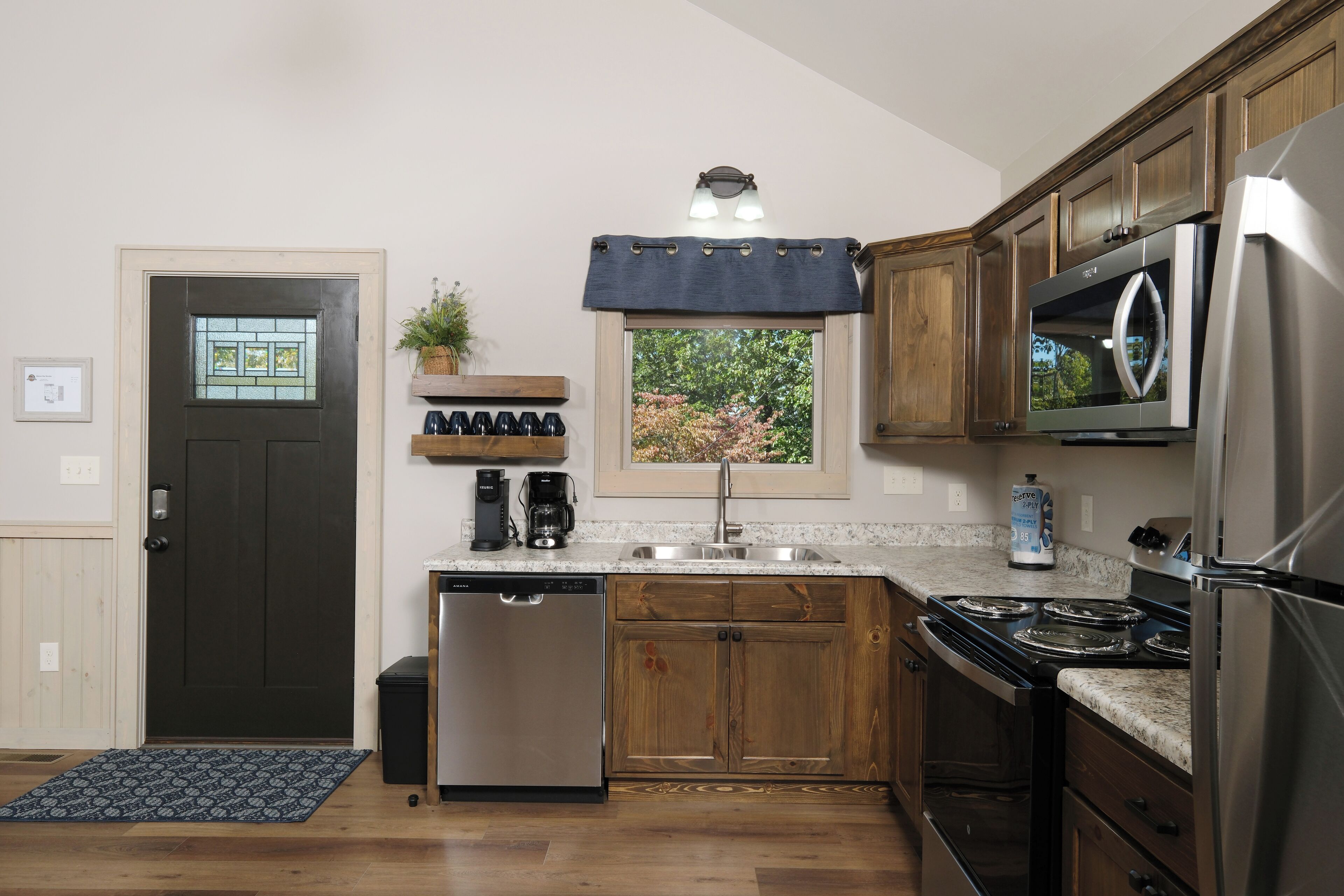A kitchen with wooden cabinets, a stainless steel refrigerator, a dishwasher, and a window with a view of trees.